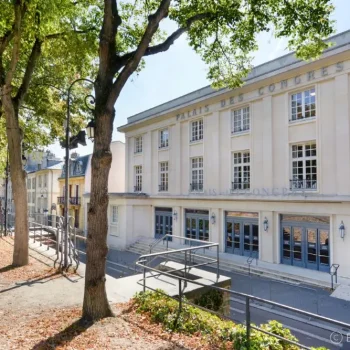 Façade du Palais des Congrès de Versailles avec architecture classique beige, grandes fenêtres, allée bordée d'arbres majestueux, ciel bleu ensoleillé