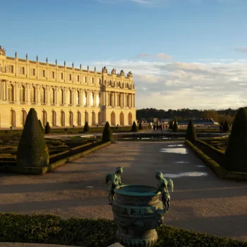 Façade majestueuse du Château de Versailles en pierre dorée, jardins à la française avec cyprès taillés, bassins classiques, allées pavées, ciel bleu dégagé en fin de journée