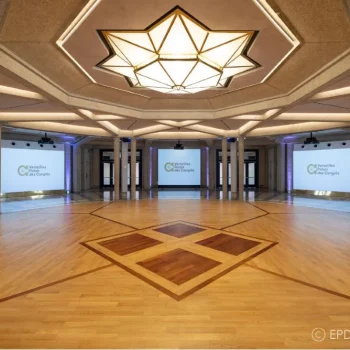 Grande salle vide avec parquet en bois clair, plafond blanc avec motifs géométriques dorés, lustre étoilé central, écrans de projection, colonnes de soutien, éclairage ambiant chaleureux